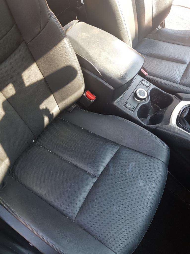 Wnętrze samochodu z widocznymi czarnymi skórzanymi fotelami z perforacją, brudnym podłokietnikiem i śladami po napojach w uchwycie.