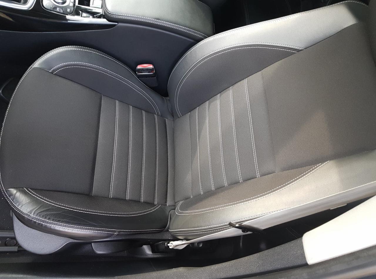 Tapicerka samochodowa w odcieniach szarości z białymi przeszyciami, widoczny fragment pasów bezpieczeństwa i detale wnętrza pojazdu.