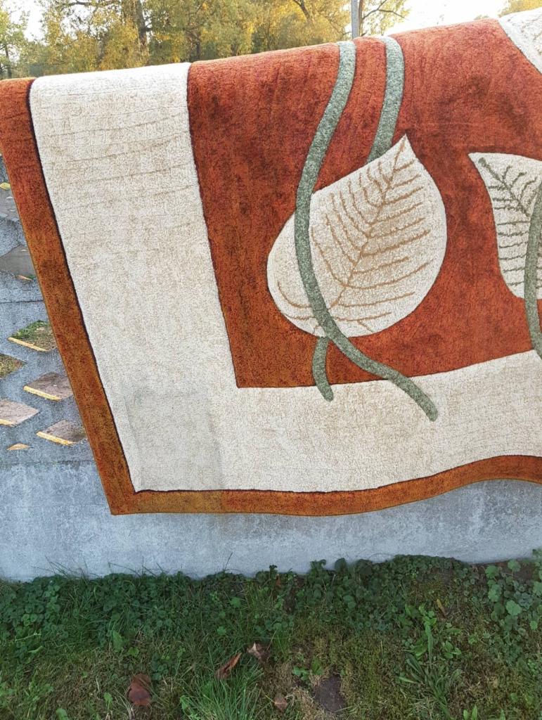 Wyprany dywan z motywem liści, suszony na zewnątrz na tle betonowego murku i trawy.