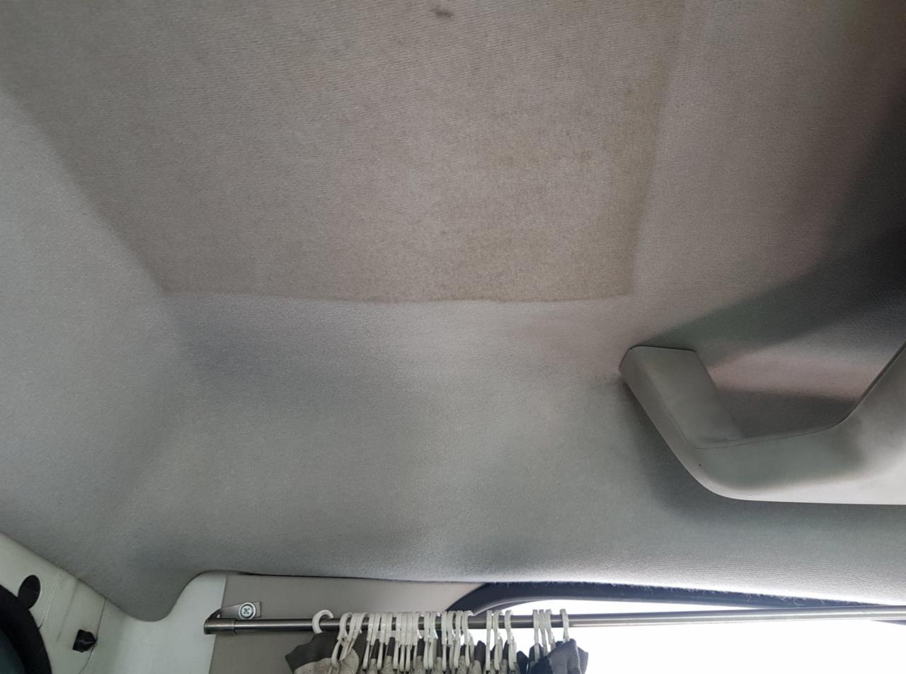 Wnętrze samochodu z widocznym sufitem tapicerowanym, fragment po czyszczeniu kontrastuje z zabrudzoną częścią, uchwyt nad drzwiami po prawej stronie, w tle wieszaki z ubraniami.