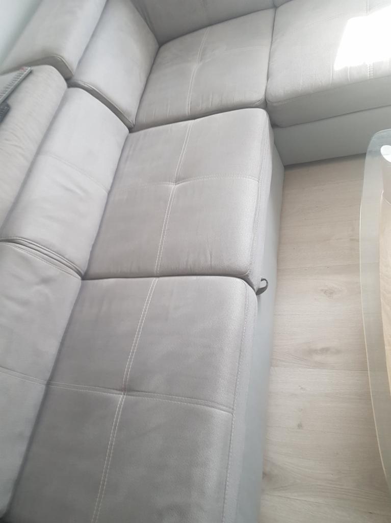 Fragment szarej, modułowej kanapy narożnej z widocznymi szwami i drewnopodobną podłogą obok szklanego stolika.