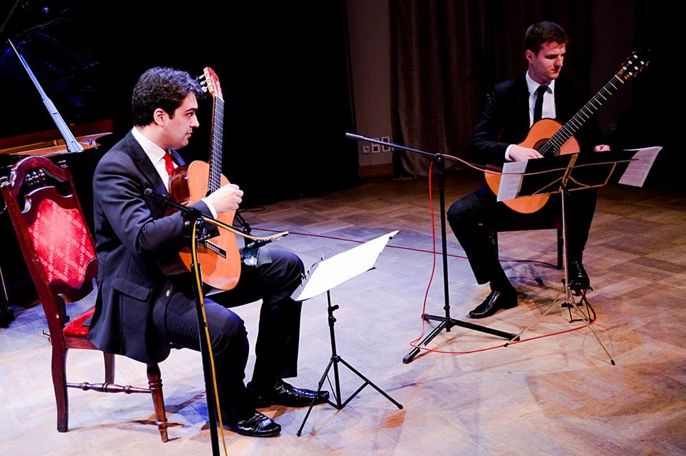 Duet gitarzystów klasycznych w czarnych garniturach, grających na scenie z drewnianą podłogą, z nutami na statywach i mikrofonami przed gitarami.