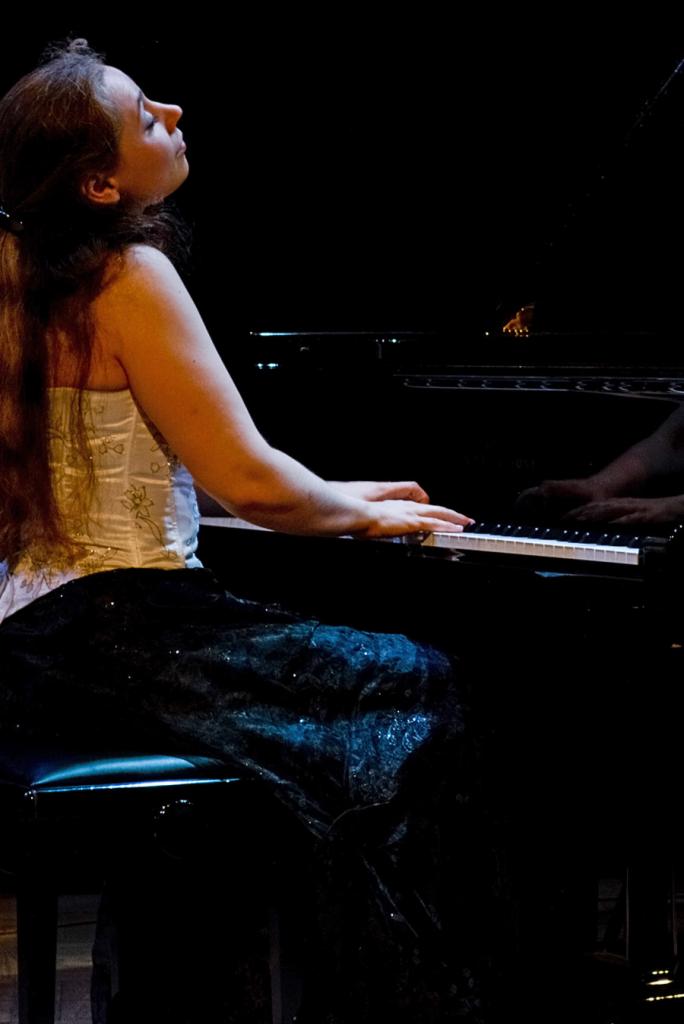 Kobieta w gorsecie i ciemnej, błyszczącej spódnicy gra na czarnym fortepianie, z zamkniętymi oczami i uniesioną głową.