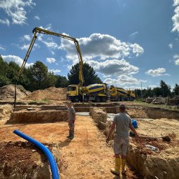 Parama Bau - Wykop pod fundamenty z widoczną pompą do betonu, dwoma betonomieszarkami w tle, oraz pracownikami w kaloszach. Niebieska rura drenarska leży na krawędzi wykopu.