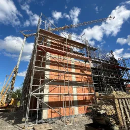 Budowa dwukondygnacyjnego budynku z czerwonej cegły i betonu, otoczonego rusztowaniami i żurawiami na tle błękitnego nieba z chmurami, widok z dołu.