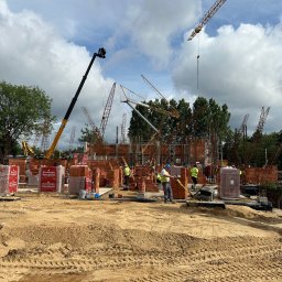 Parama Bau - Budowa domu wielorodzinnego w toku: widoczne fundamenty, ściany z czerwonej cegły Porotherm, stalowe zbrojenia, pracownicy w kamizelkach odblaskowych, dźwigi budowlane w tle, teren przygotowany...