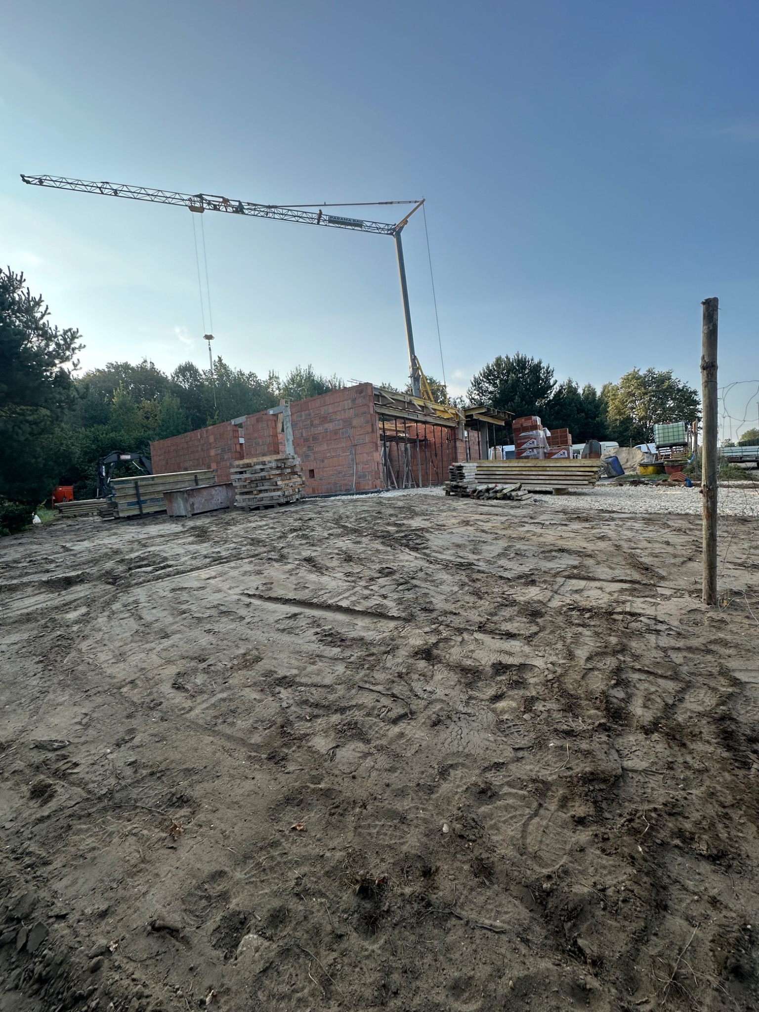 Wznoszenie murów z czerwonej cegły na placu budowy w Bodzanowicach, widoczny żuraw budowlany, palety z materiałami, wykopana ziemia i częściowo zrealizowana konstrukcja budynku.