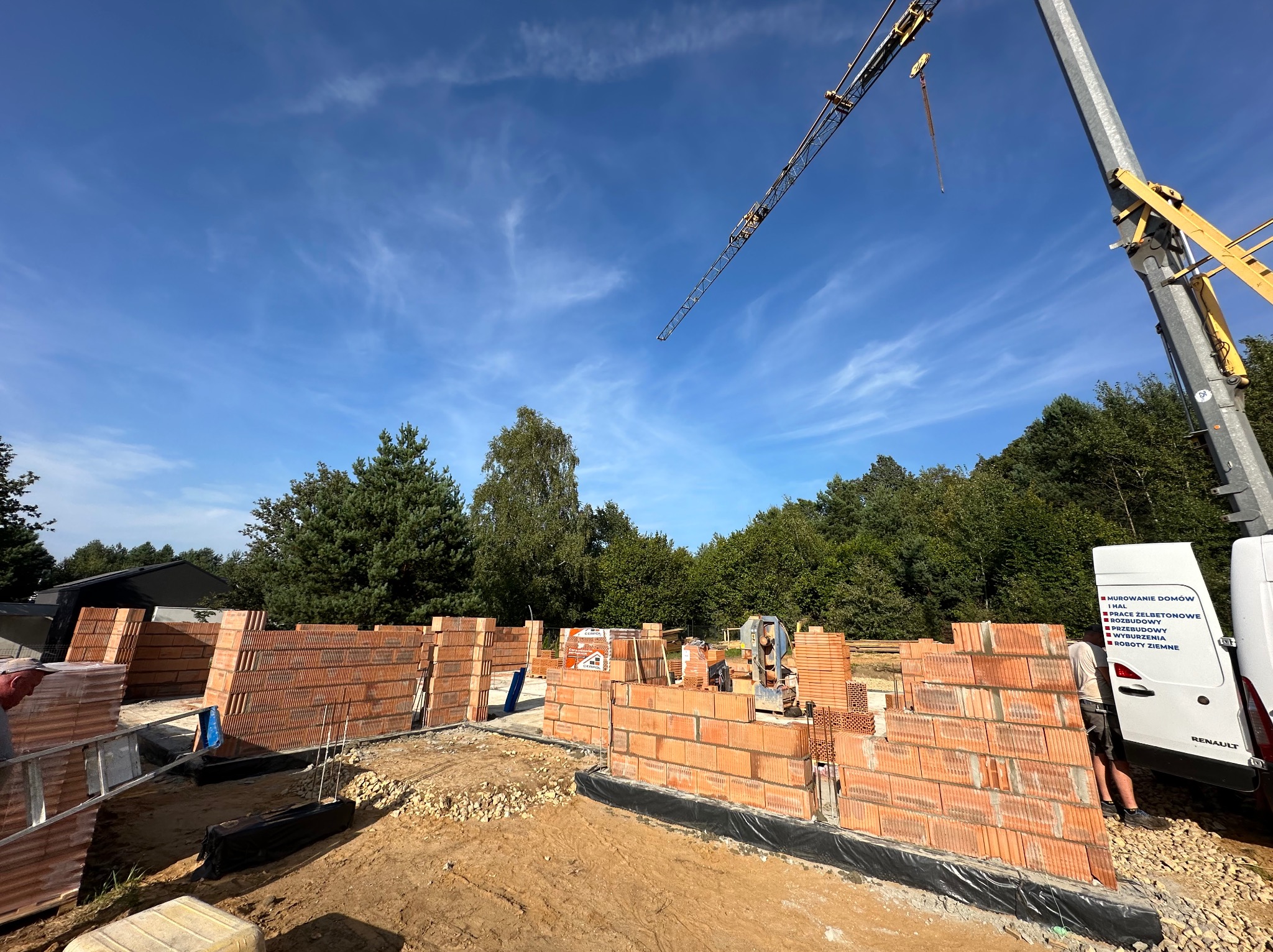Wznoszenie ścian fundamentowych z czerwonej cegły ceramicznej na budowie, widoczny dźwig budowlany i częściowo biały samochód dostawczy z reklamą firmy budowlanej.