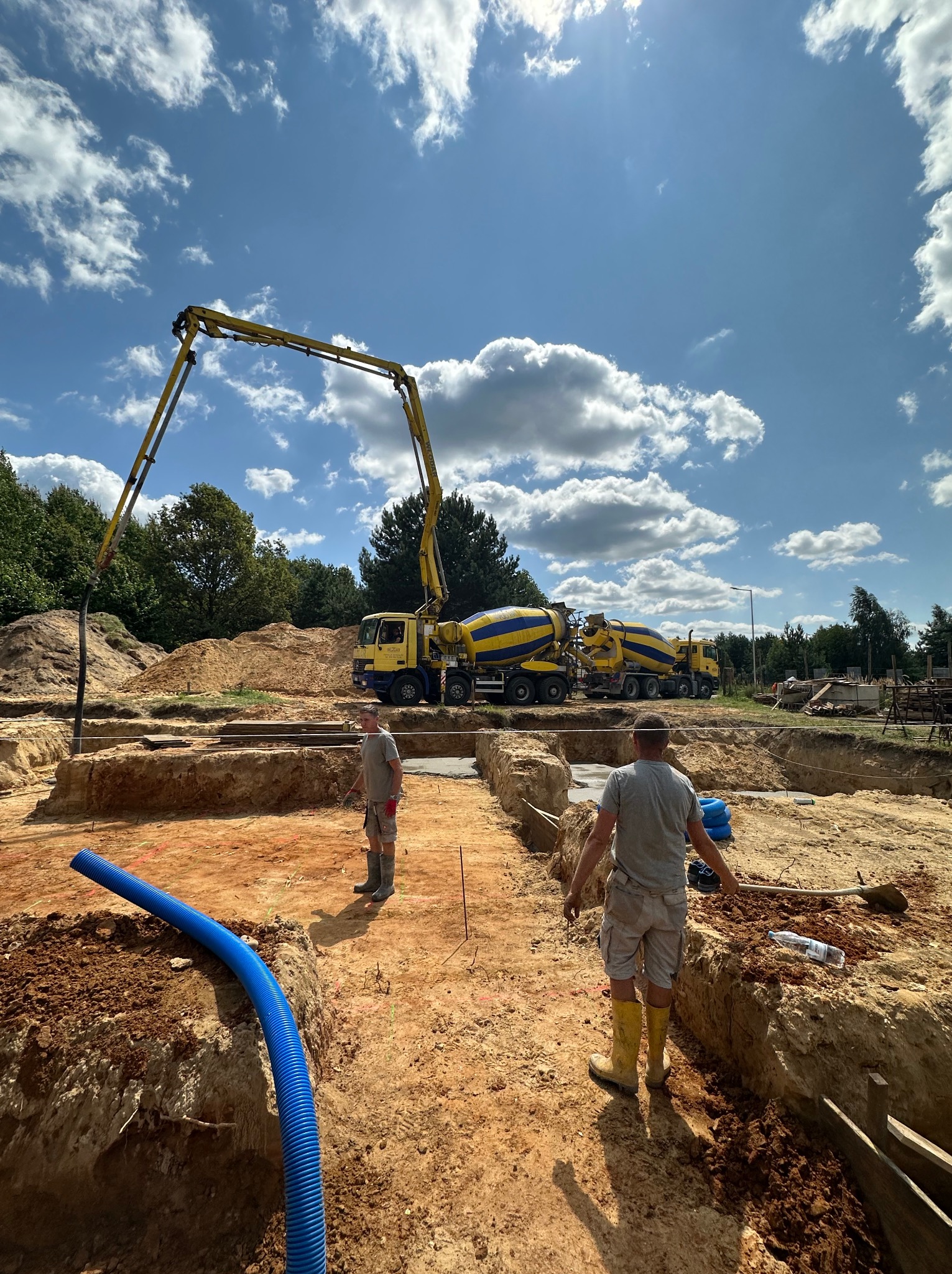 Wykop pod fundamenty z widoczną pompą do betonu, dwoma betonomieszarkami w tle, oraz pracownikami w kaloszach. Niebieska rura drenarska leży na krawędzi wykopu.