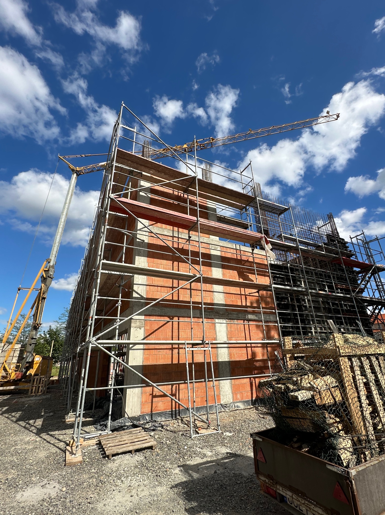 Budowa dwukondygnacyjnego budynku z czerwonej cegły i betonu, otoczonego rusztowaniami i żurawiami na tle błękitnego nieba z chmurami, widok z dołu.