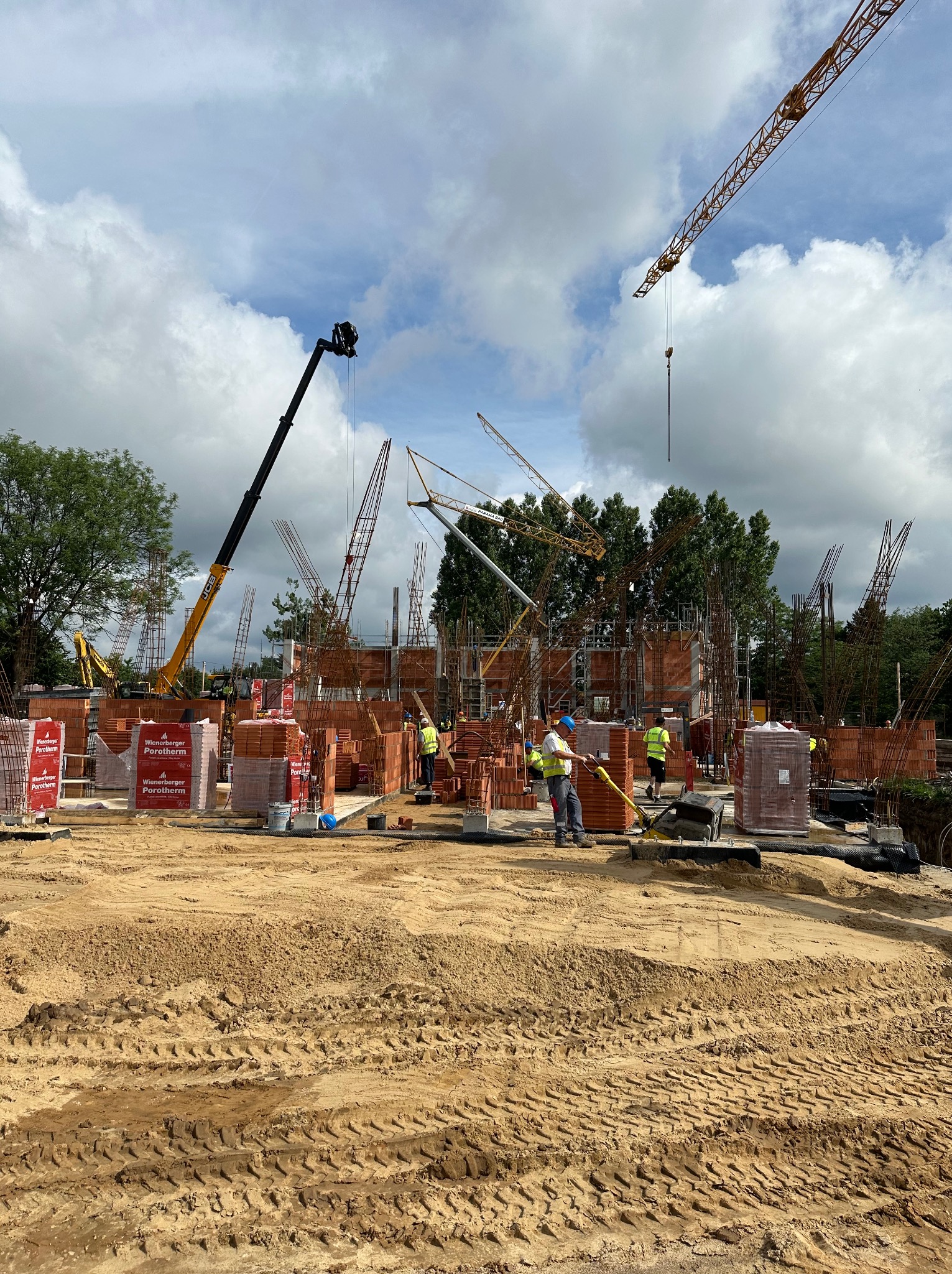 Budowa domu wielorodzinnego w toku: widoczne fundamenty, ściany z czerwonej cegły Porotherm, stalowe zbrojenia, pracownicy w kamizelkach odblaskowych, dźwigi budowlane w tle, teren przygotowany...