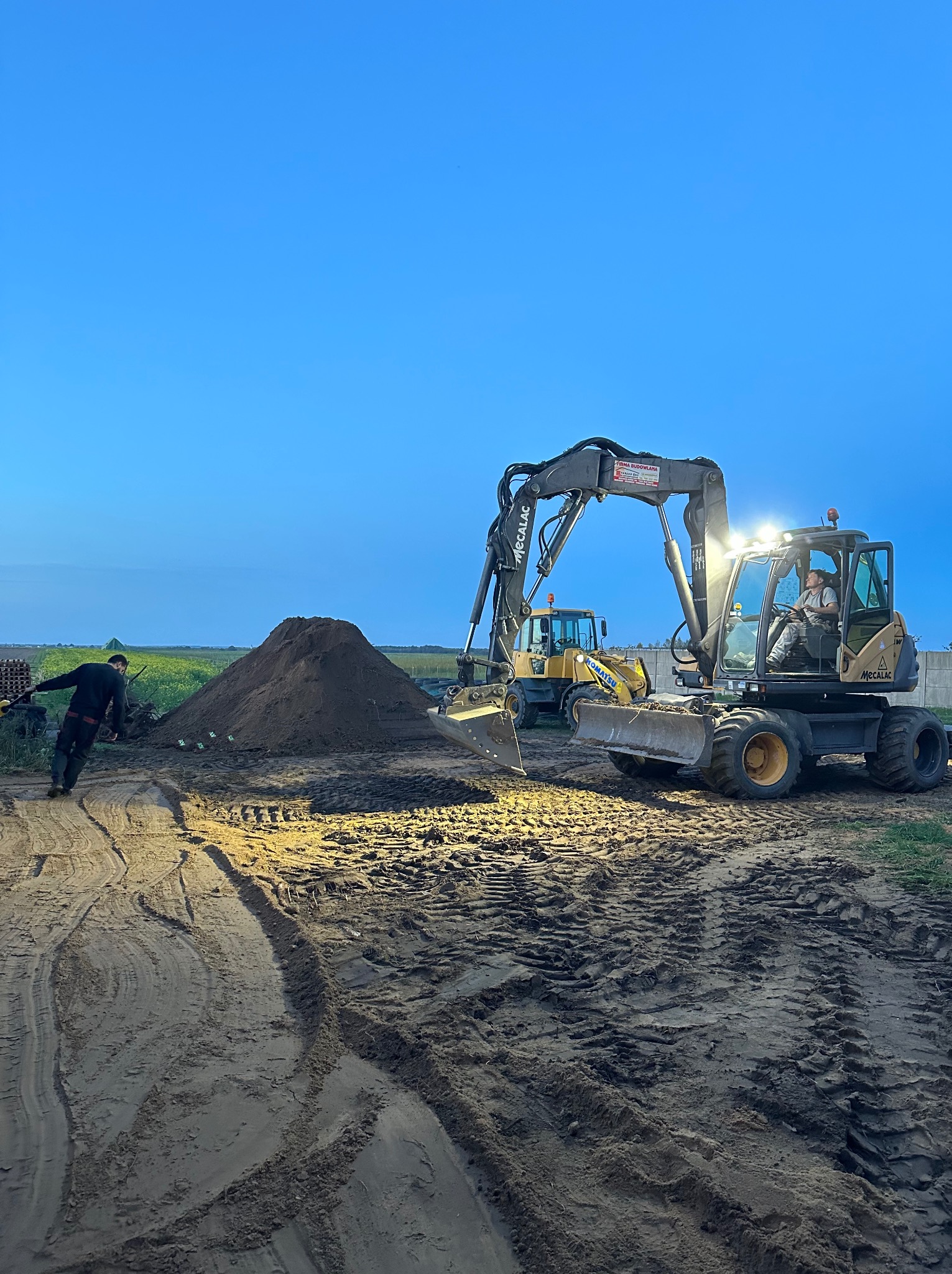 Operator w kabinie koparki Mecalac z włączonymi światłami, obok sterta ziemi i pracownik z łopatą, w tle druga koparka Komatsu, teren budowy o zmierzchu.