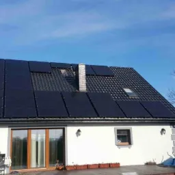 Dom jednorodzinny z panelami fotowoltaicznymi na dachu, widoczne okna dachowe i komin, jasne niebo w tle.