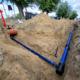 Głęboki wykop z ułożonymi niebieskimi rurami wodociągowymi, widoczne złączki i narzędzie wbite w ziemię, w tle pryzma piasku i fragment zabudowań.