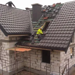 Montaż ciemnej dachówki na nowym domu w stanie surowym, dekarz w żółtej kurtce układa dachówkę przy oknie dachowym, drugi dekarz pracuje na sąsiedniej połaci dachu.