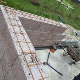 Wznoszenie ściany z bloczków z keramzytu zbrojonej siatką stalową, widoczne narzędzia budowlane i taczka z zaprawą w otoczeniu trawy i ogrodzenia.