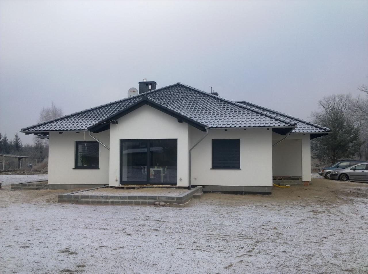 Nowoczesny, parterowy dom jednorodzinny z ciemną dachówką i stolarką okienną, w trakcie wykańczania terenu wokół budynku, z lekkim śniegiem na dachu i ziemi.