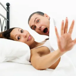 Kobieta w łóżku z wyciągniętą dłonią, zaskoczona przez mężczyznę wyłaniającego się spod kołdry z otwartymi ustami.