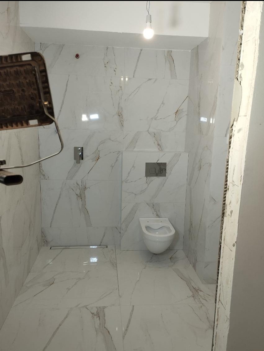 Biała łazienka z marmurowymi płytkami w trakcie remontu, widoczny fragment lampy budowlanej, zamontowana toaleta podwieszana i odpływ liniowy w podłodze.