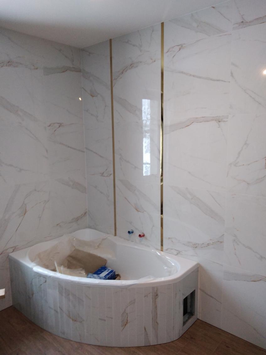 Wykończona łazienka z wanną narożną obudowaną płytkami imitującymi marmur, ściany wyłożone dużymi płytami z motywem marmuru, złote listwy dekoracyjne, widoczne przyłącza wodne.