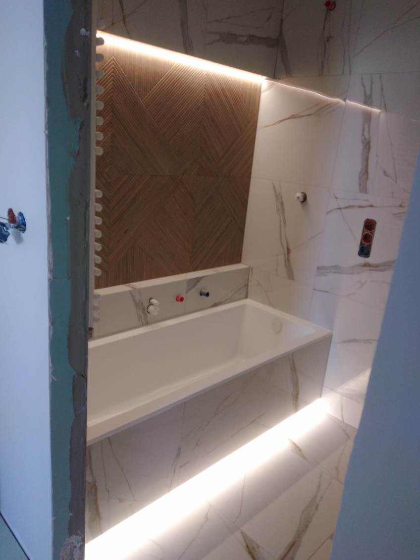 Nowoczesna łazienka w trakcie wykańczania: biała wanna obudowana marmurowymi płytkami, drewniana mozaika na ścianie podświetlona listwą LED, widoczne elementy montażowe na ścianie i niezakończone...