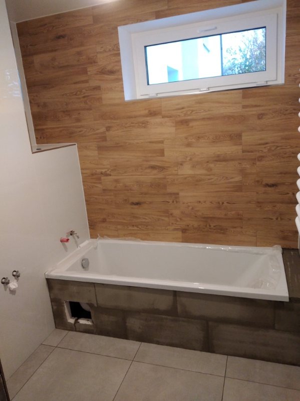 Biała wanna obudowana szarą mozaiką, ściany wyłożone panelami imitującymi drewno, widoczne niedokończone prace wykończeniowe, surowy stan.