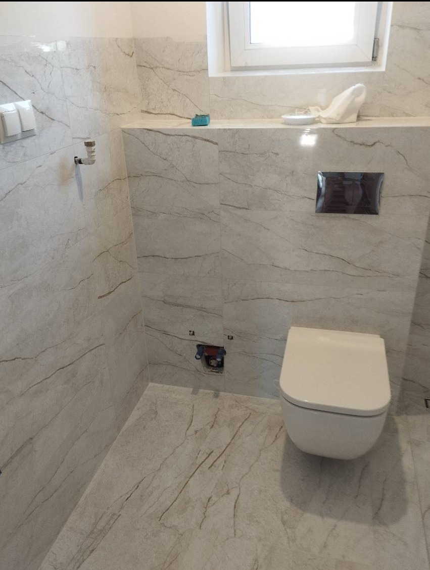 Nowoczesna łazienka z płytkami imitującymi marmur, wisząca toaleta, minimalistyczny przycisk spłuczki i widoczne przyłącze kanalizacyjne na ścianie.