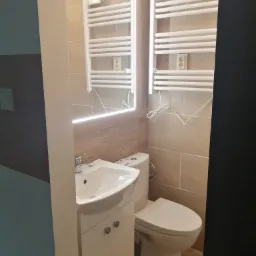 Nowoczesna łazienka z wiszącą toaletą, białą umywalką z szafką, podświetlanym lustrem i grzejnikiem drabinkowym na ścianie z beżowych płytek.