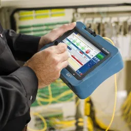 Technik używa testera MaxTester do diagnostyki światłowodów w szafie telekomunikacyjnej, wyświetlając wykresy i dane pomiarowe.