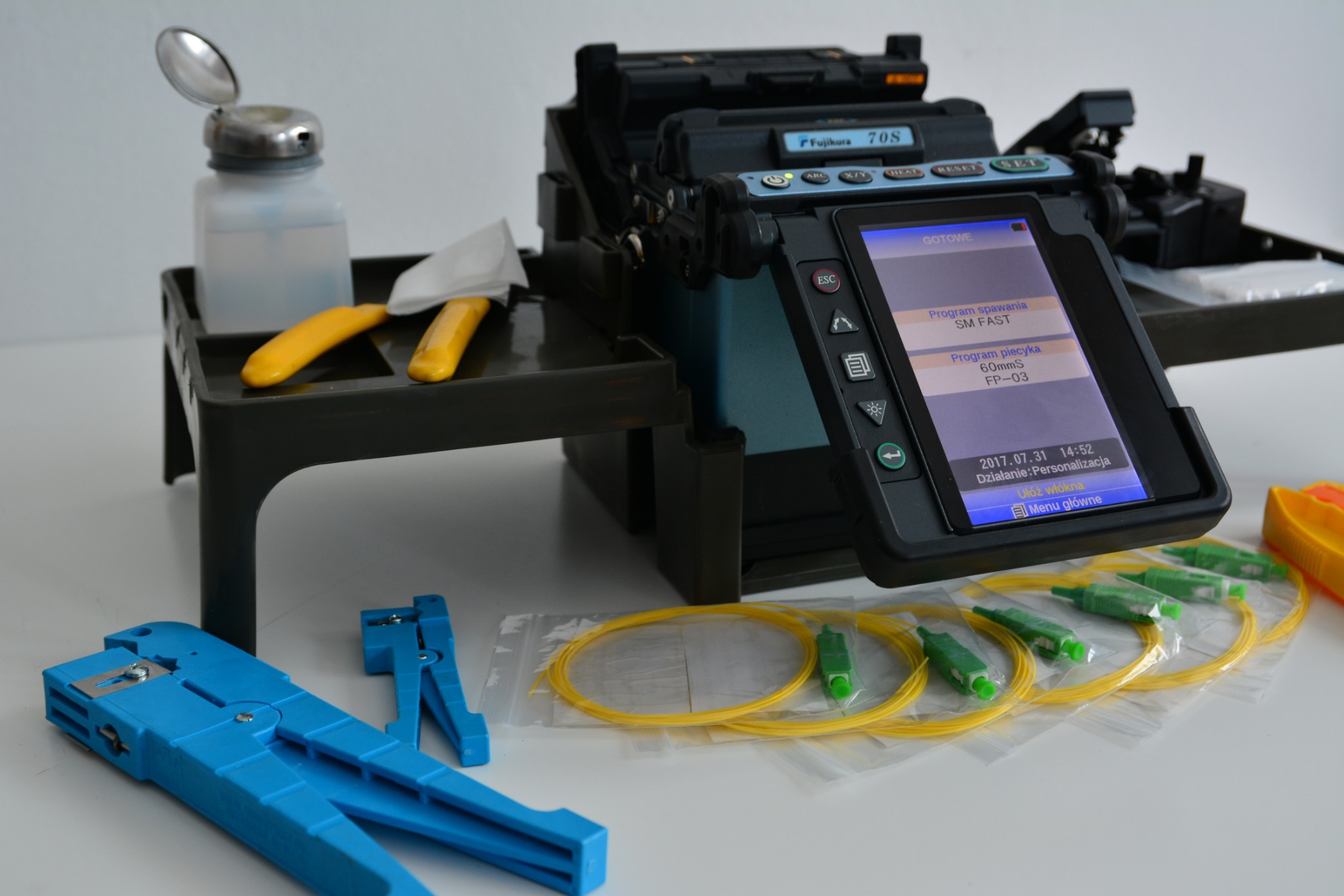 Zgrzewarka światłowodowa Fujikura 70S z akcesoriami: czyścikiem, pojemnikiem na alkohol izopropylowy, stripperem światłowodowym oraz złączkami światłowodowymi SC/APC, na białym tle.