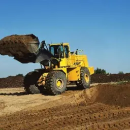 Żółta ładowarka kołowa z pełną łyżką ziemi na tle błękitnego nieba i hałd ziemi, widoczne ślady opon na ziemi.