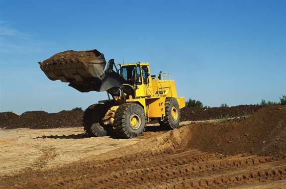 Żółta ładowarka kołowa z pełną łyżką ziemi na tle błękitnego nieba i hałd ziemi, widoczne ślady opon na ziemi.