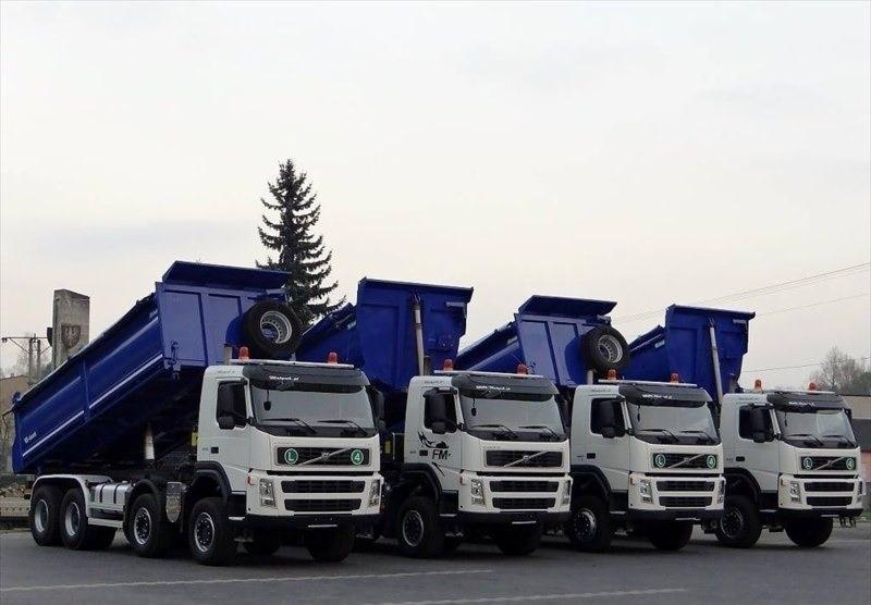 Flota białych wywrotek Volvo z niebieskimi skrzyniami załadunkowymi, zaparkowana w Magdalence, z choinką w tle.