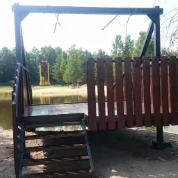 Stalowa platforma widokowa z drewnianą balustradą i schodami, zawieszona nad brzegiem jeziora, z linami podtrzymującymi konstrukcję, w tle las i wieża obserwacyjna.