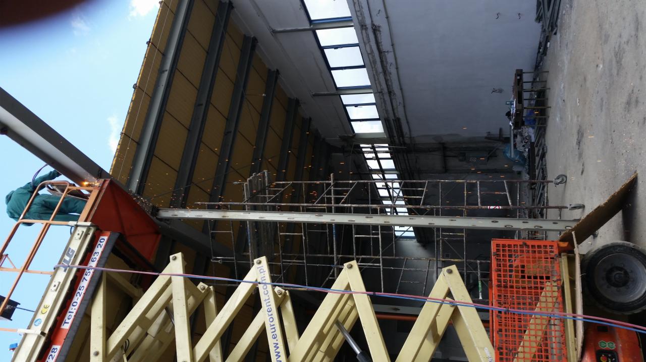 Spawanie konstrukcji stalowej hali z podnośnika koszowego, widoczne iskry i elementy konstrukcyjne dachu hali.