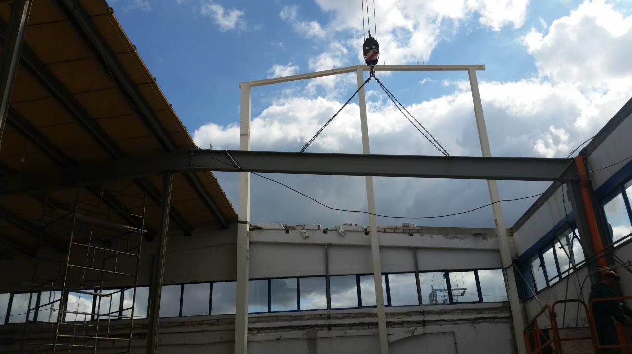 Montaż stalowej ramy hali przemysłowej z użyciem dźwigu na tle pochmurnego nieba; widoczne elementy konstrukcyjne i fragment budynku.