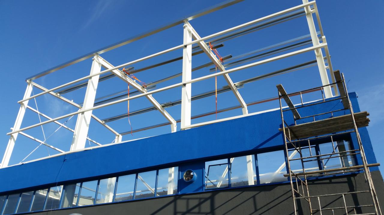 Konstrukcja stalowa hali w trakcie budowy, widoczne białe elementy szkieletu na tle błękitnego nieba, niebieska ściana budynku i rusztowanie z drewnianymi deskami.