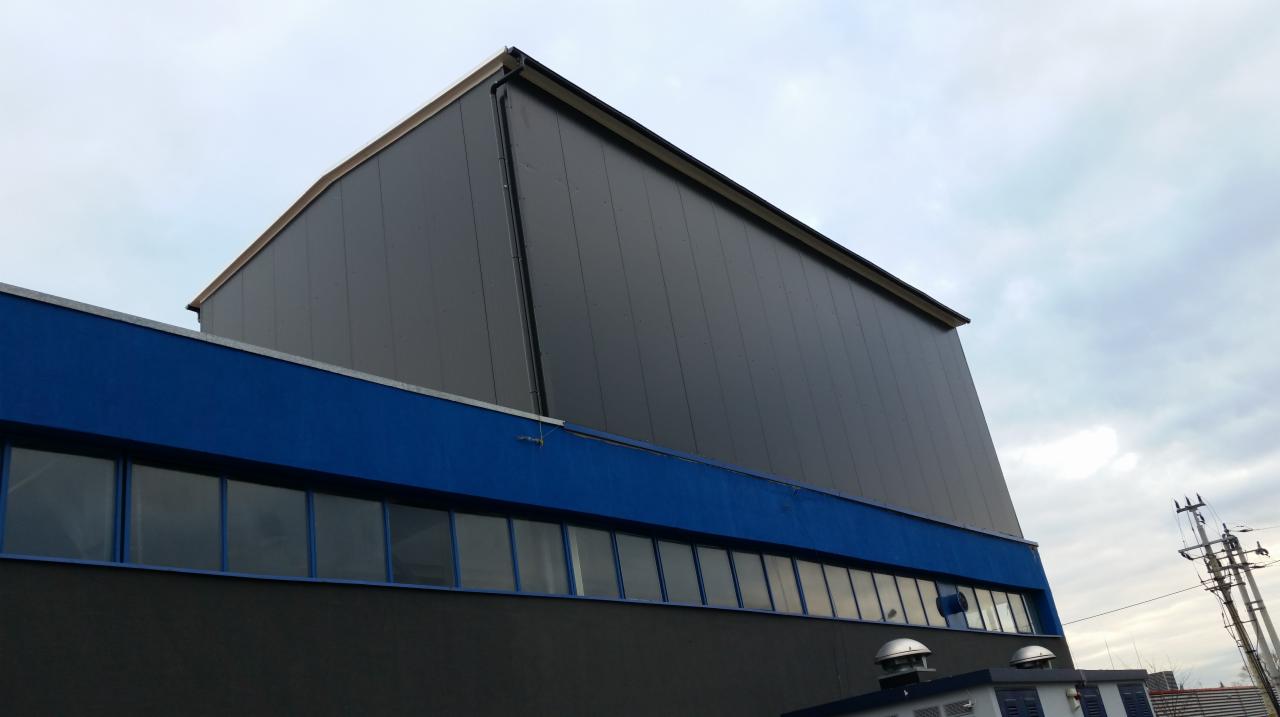 Zewnętrzny widok hali przemysłowej z szarej płyty warstwowej, z niebieskim pasem okiennym w dolnej części, uchwycony pod kątem z dołu w pochmurny dzień.