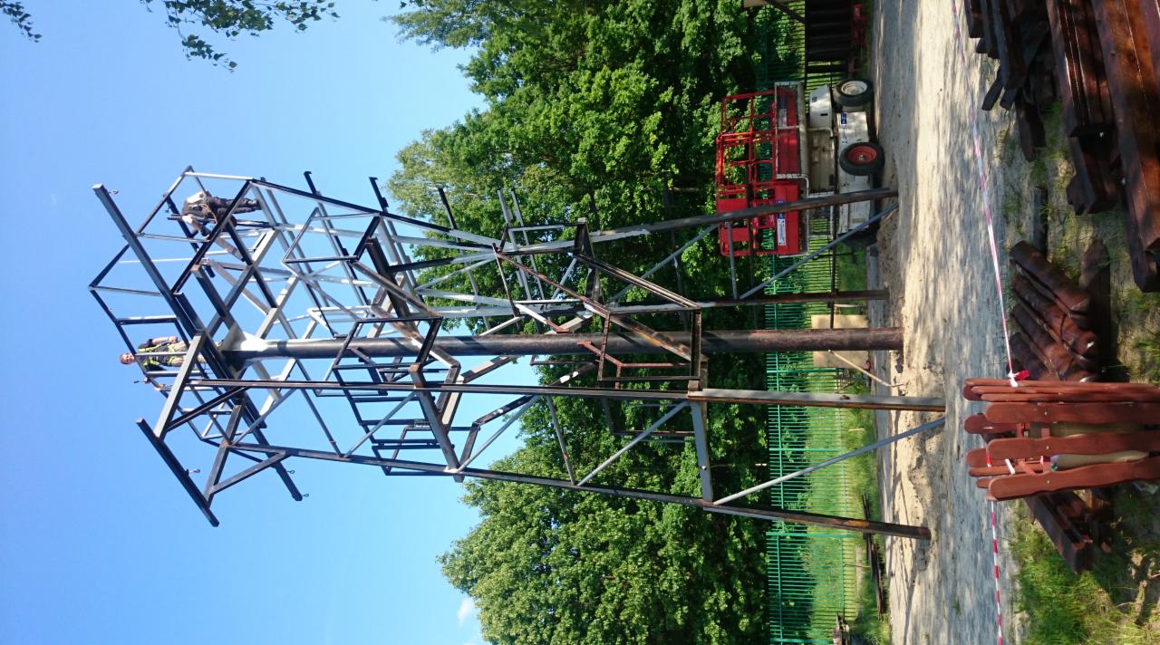 Montaż ażurowej konstrukcji stalowej przez dwóch pracowników na wysokości, z użyciem podnośnika koszowego, widok z dołu na tle błękitnego nieba i zieleni drzew.