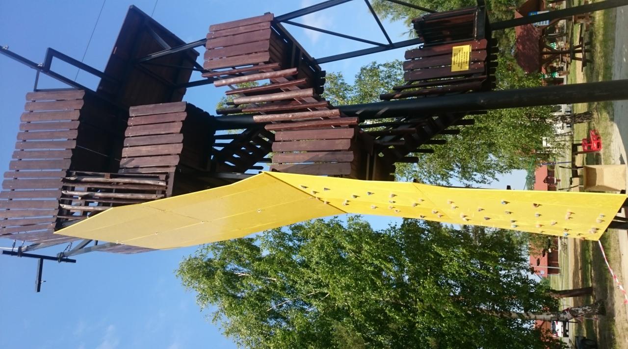 Plac zabaw z drewnianą konstrukcją i żółtą ścianką wspinaczkową na tle drzew i nieba.