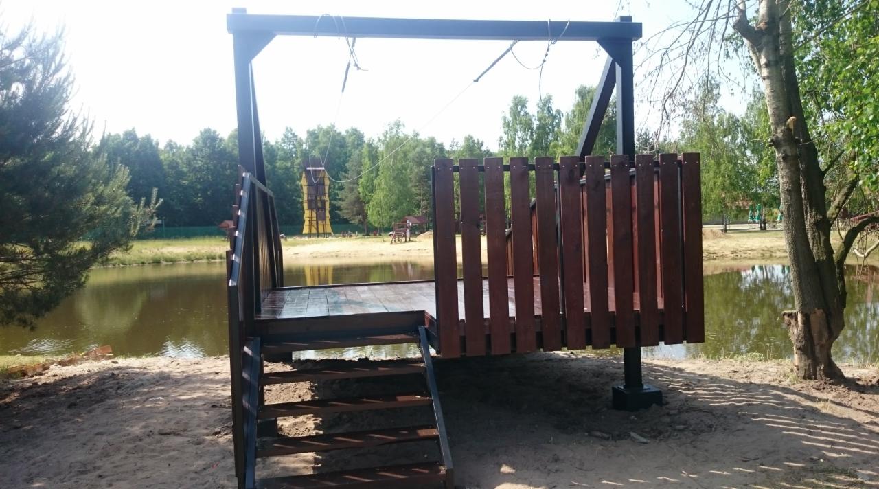 Stalowa platforma widokowa z drewnianą balustradą i schodami, zawieszona nad brzegiem jeziora, z linami podtrzymującymi konstrukcję, w tle las i wieża obserwacyjna.