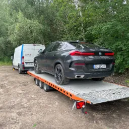 Czarny BMW X6 na lawecie holowanej przez biały bus Peugeot. Transport pojazdu w otoczeniu zieleni.