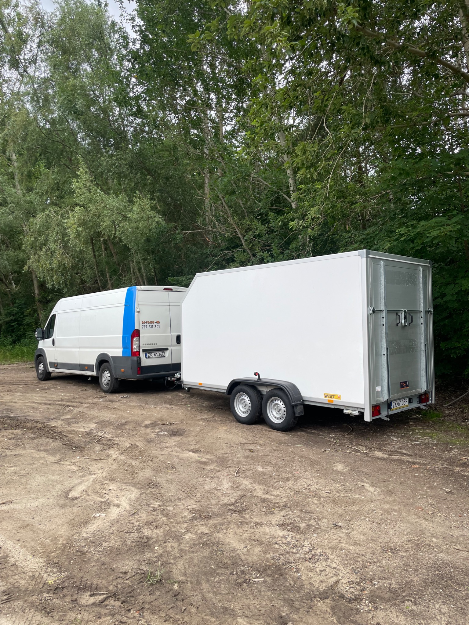 Biały bus z przyczepą na piaszczystym terenie, otoczony zielenią. Bus z niebieskim pasem i numerem telefonu. Przyczepa z podwójnymi drzwiami i tablicą rejestracyjną.