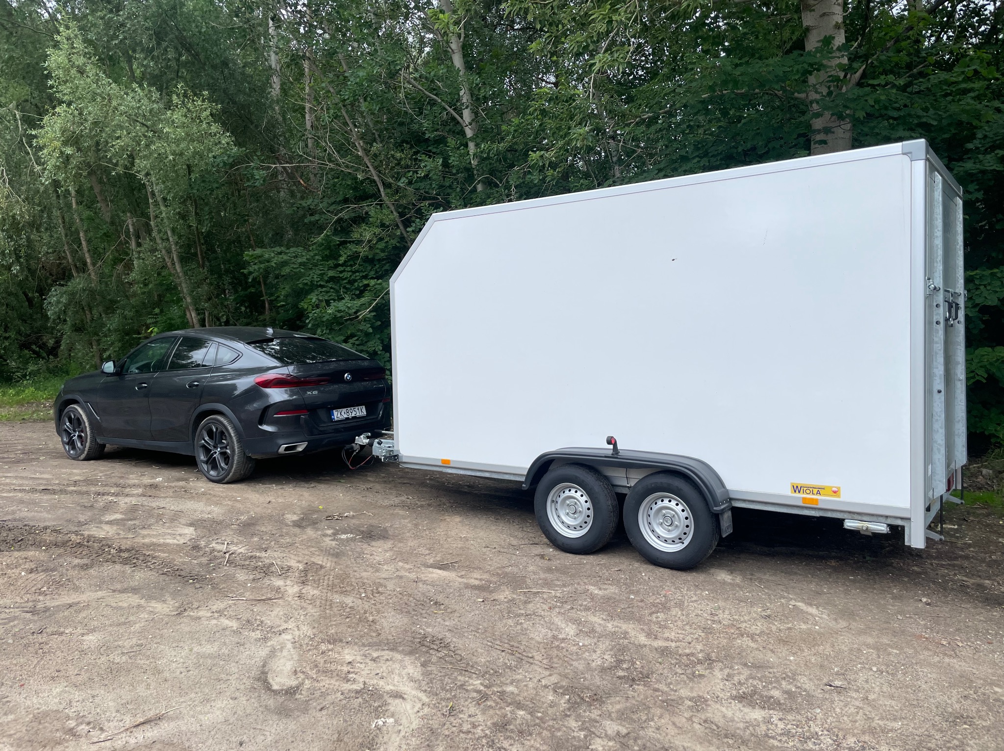 Ciemny SUV BMW X6 ciągnie białą, zamkniętą przyczepę transportową na leśnej drodze, gotowy do transportu. Widoczny hak holowniczy i tablica rejestracyjna.