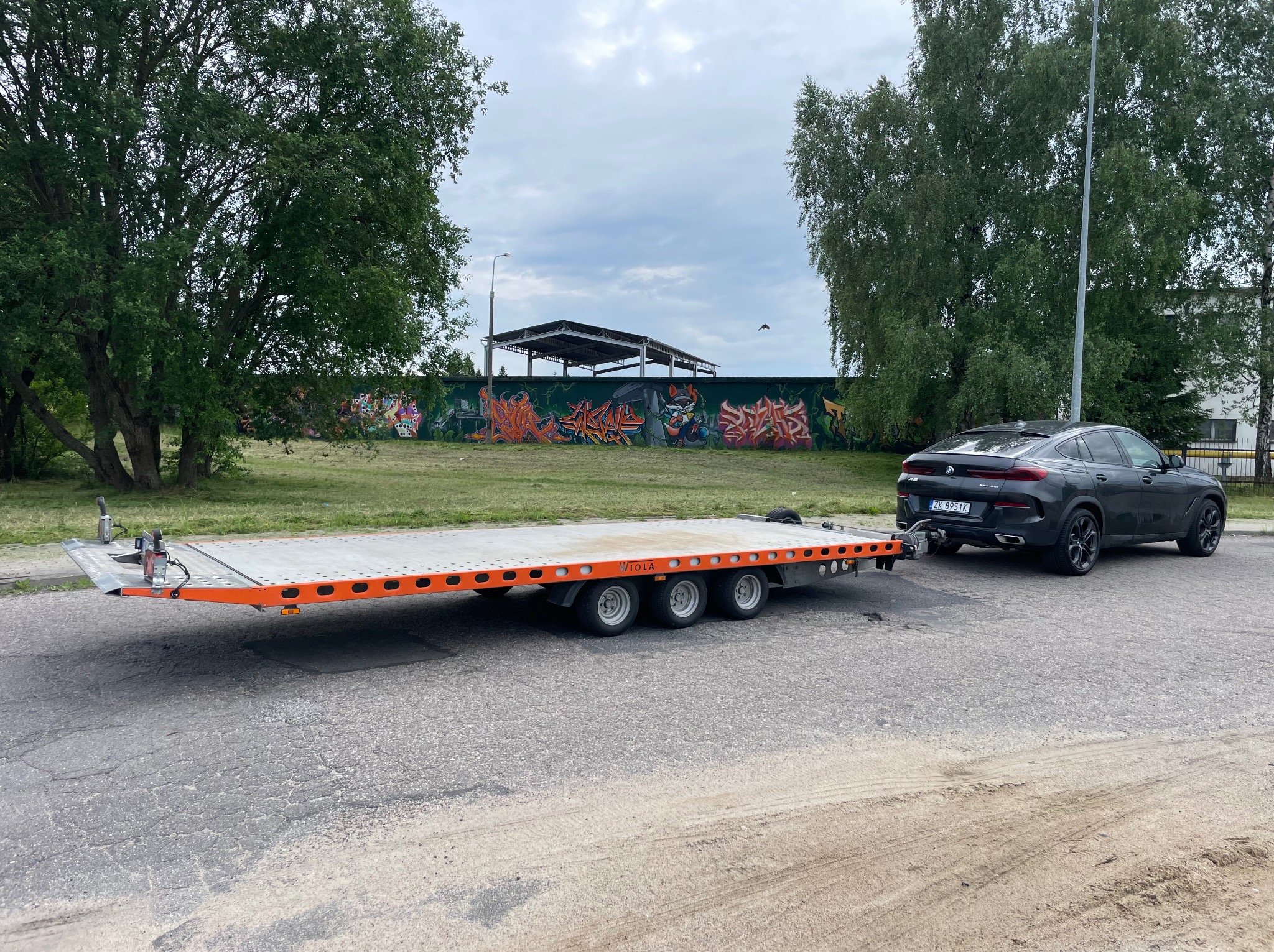 Czarny SUV ciągnie niskopodwoziową przyczepę z pomarańczowymi akcentami na tle graffiti. Widoczne drzewa i pochmurne niebo.