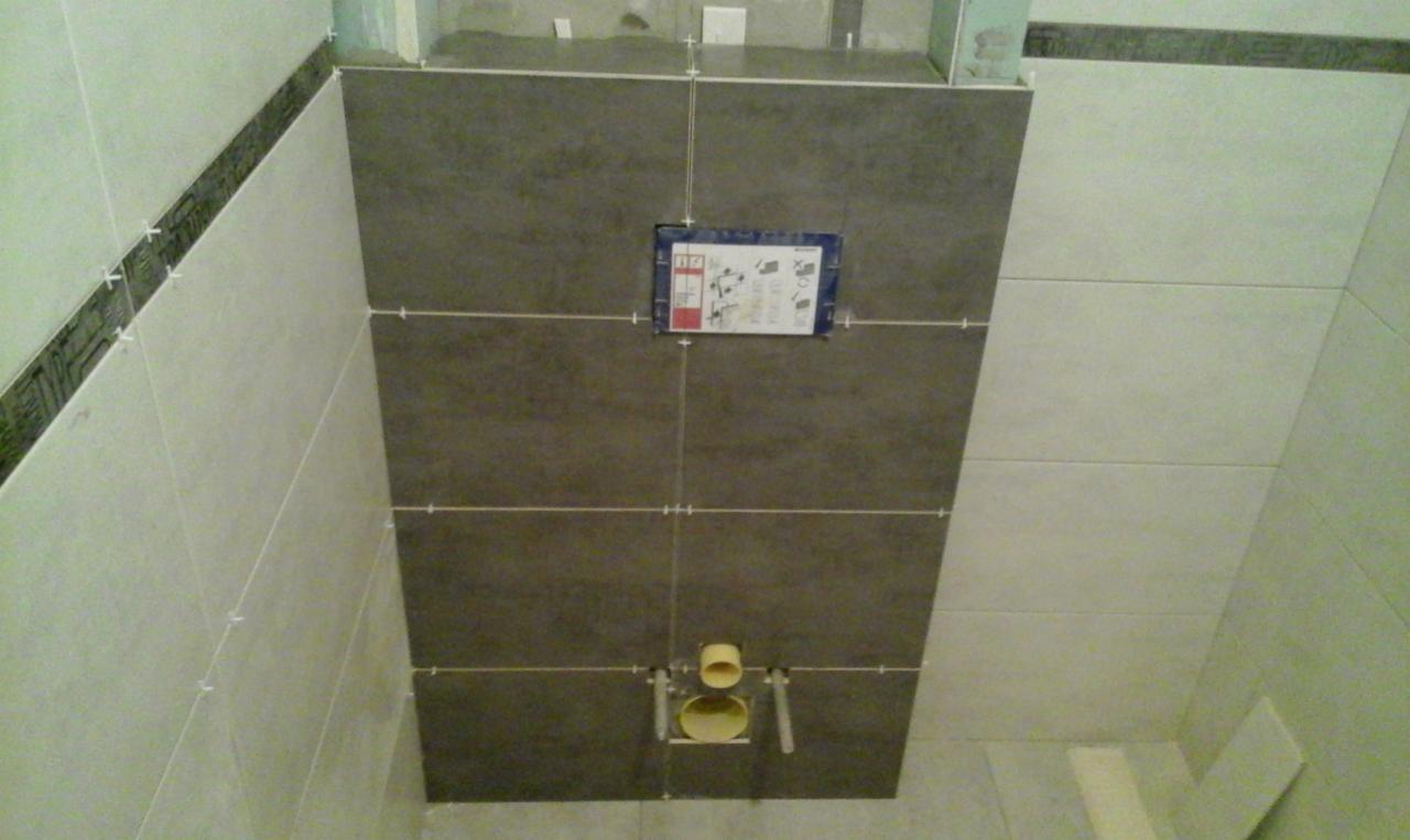 Montaż płytek ceramicznych w łazience, widoczne szare i białe płytki, krzyżyki dystansowe, oraz elementy instalacji podtynkowej WC.