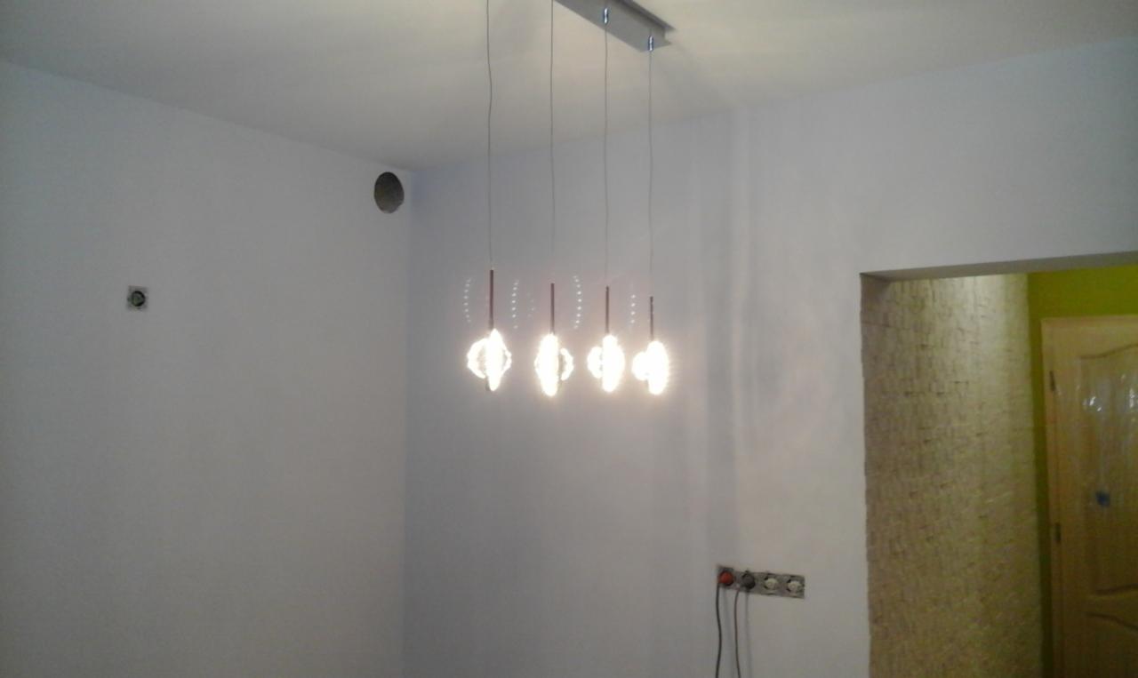 Nowoczesna, czteropunktowa lampa wisząca z dekoracyjnymi kloszami oświetla surowe, białe wnętrze w trakcie wykańczania, widoczne puszki elektryczne i częściowo wykończone wejście do pomieszczenia...