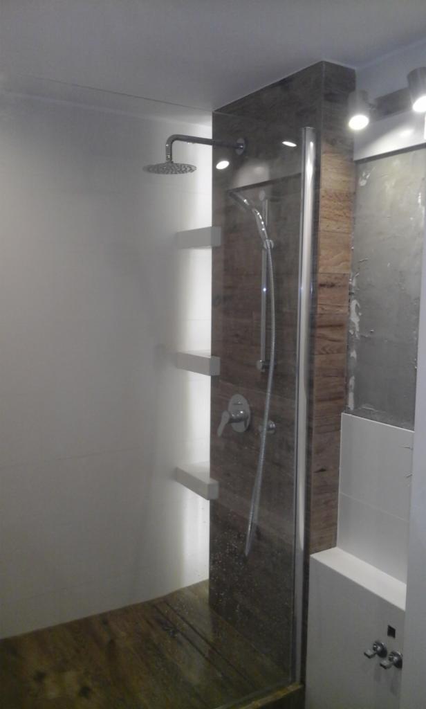 Nowoczesna kabina prysznicowa z deszczownicą i drewnopodobnymi płytkami, widoczne półki na ścianie i szklane drzwi z chromowanym wykończeniem.