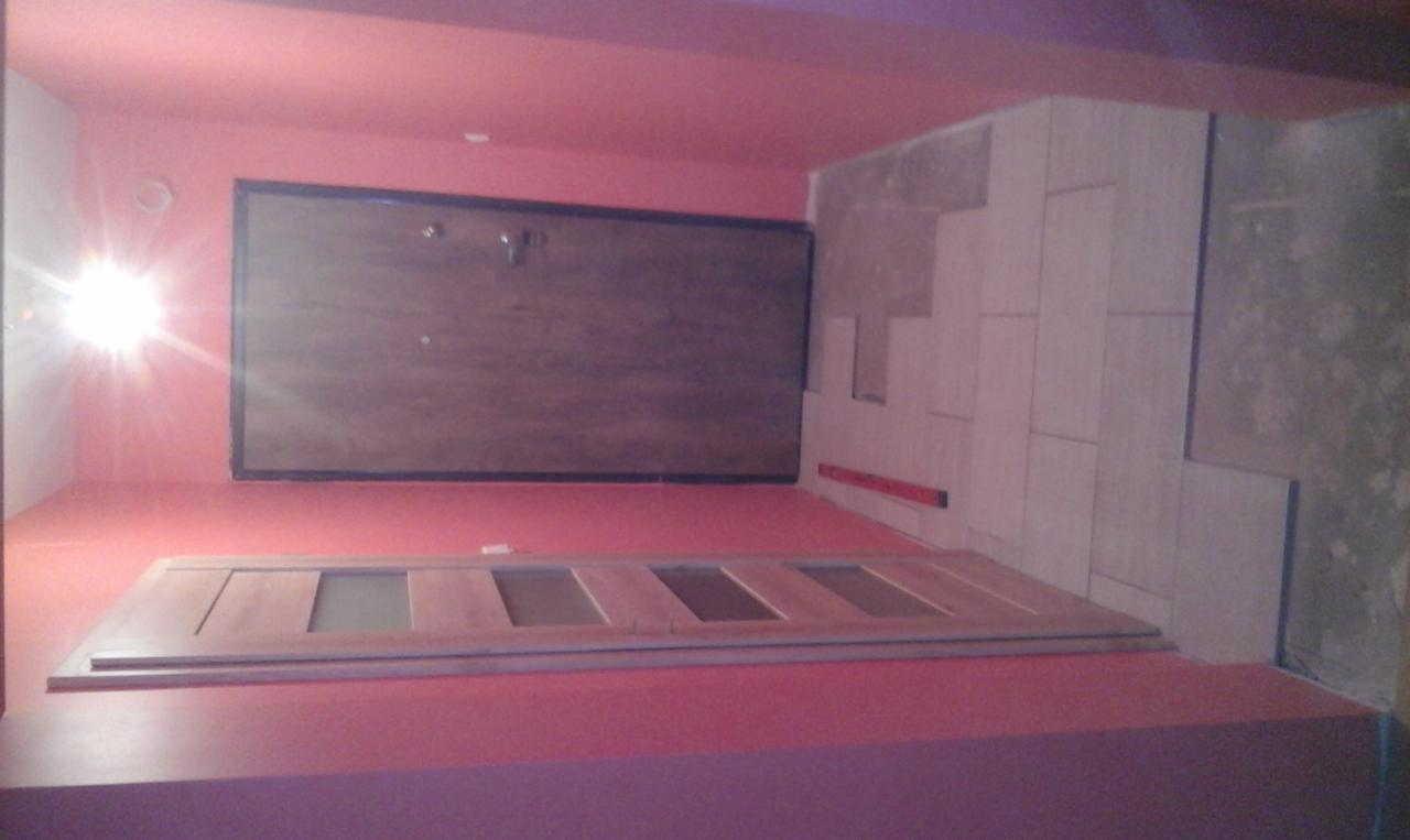 Łazienka w trakcie wykończenia: częściowo ułożone płytki na ścianie, poziomica i drzwi z szybami leżące na podłodze, ściany pomalowane na różowo.