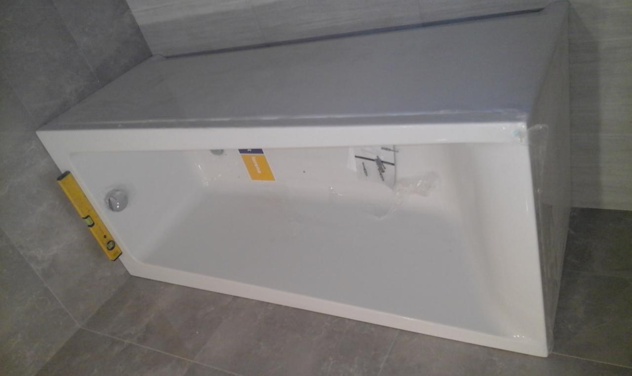 Nowa, biała wanna akrylowa, częściowo owinięta folią ochronną, ustawiona na szarych płytkach podłogowych, z poziomnicą przyłożoną do boku, gotowa do obudowy.